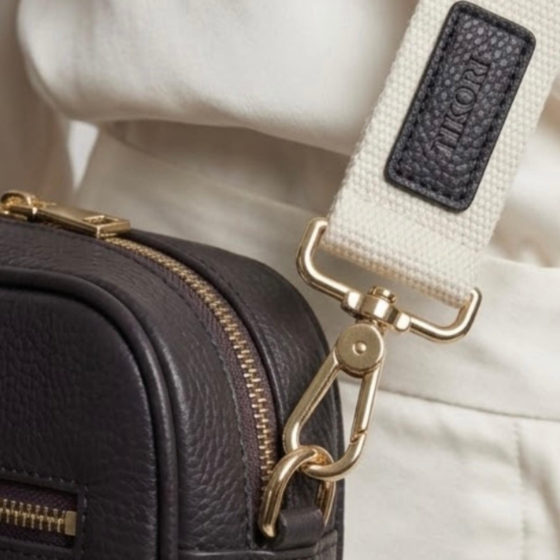 Classic elegance with practicality and versatility in mind. TIKORI classic dark brown by TIKORI london. Genuine leather crossbody bag. Model in ivory cloths wears the crossbody. Close up details of the golden hardware and canvas strap showing in picture.