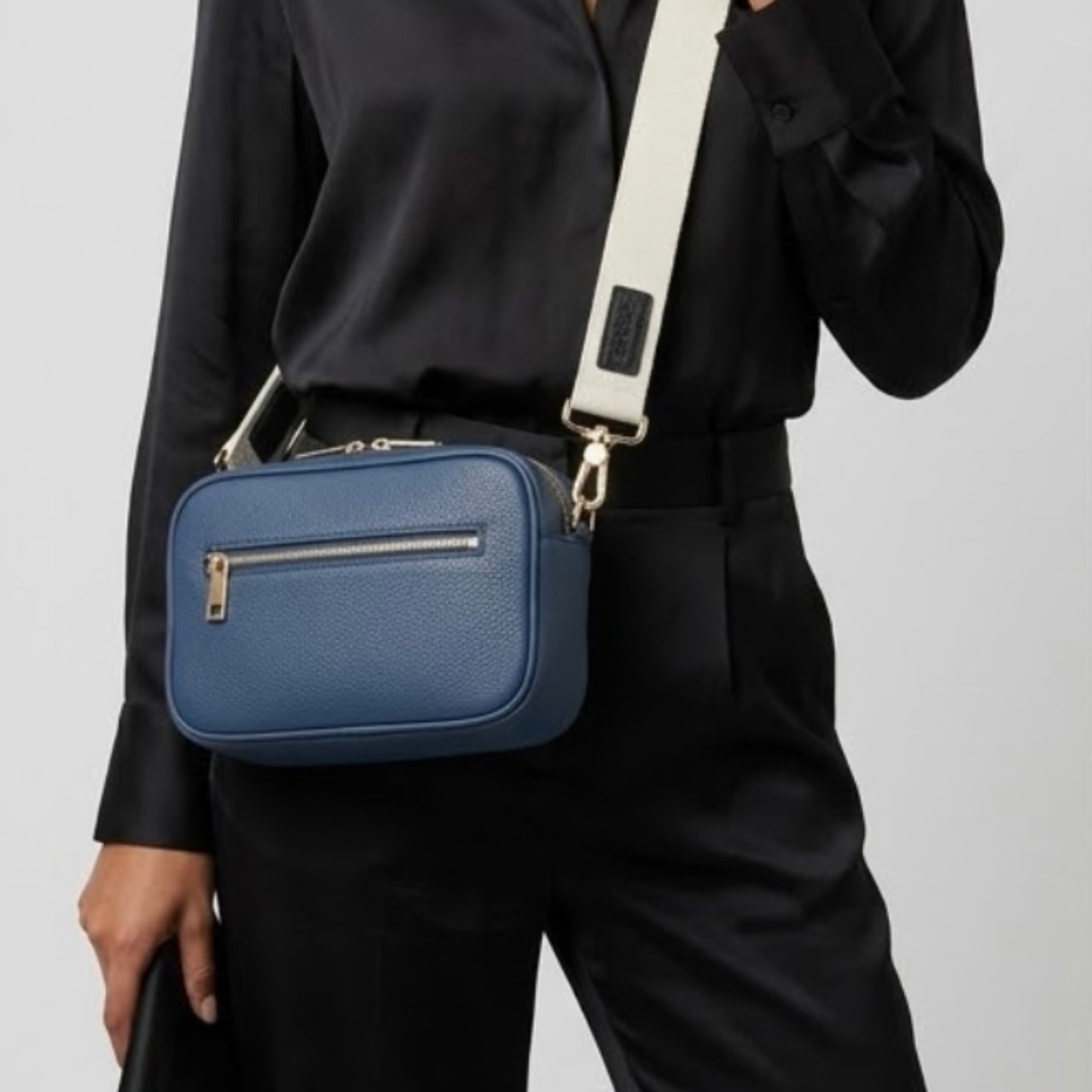 Classic elegance with practicality and versatility in mind. TIKORI classic Navy by TIKORI london. Genuine Leather cross body bag. Model in black silk cloths wears the crossbody. Front profile of the bag showing with golden tone hardware and front pocket details with zipper, in Fully white background.