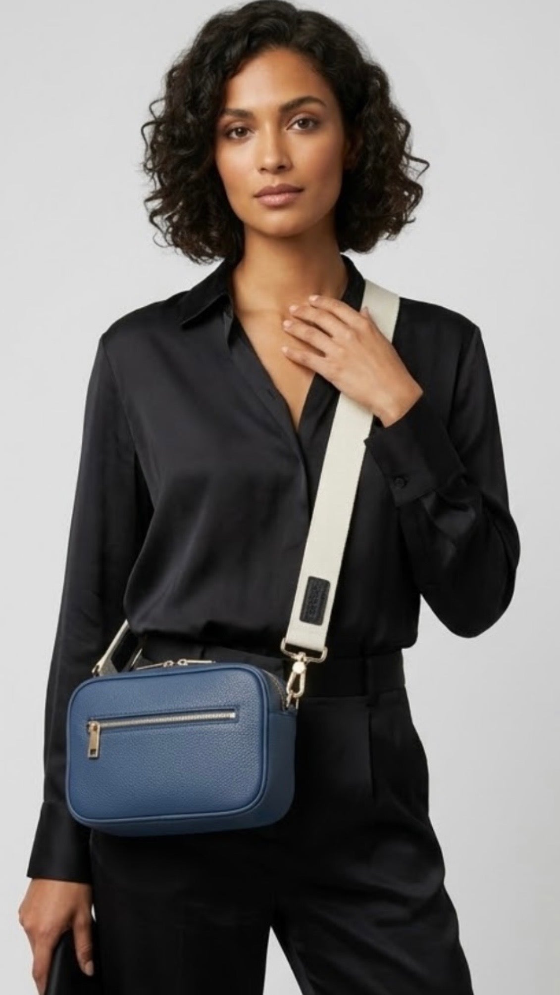 Classic elegance with practicality and versatility in mind. TIKORI classic Navy by TIKORI london. Genuine Leather cross body bag. Model in black silk cloths wears the crossbody. Front profile of the bag showing with golden tone hardware and front pocket details with zipper, in Fully white background.