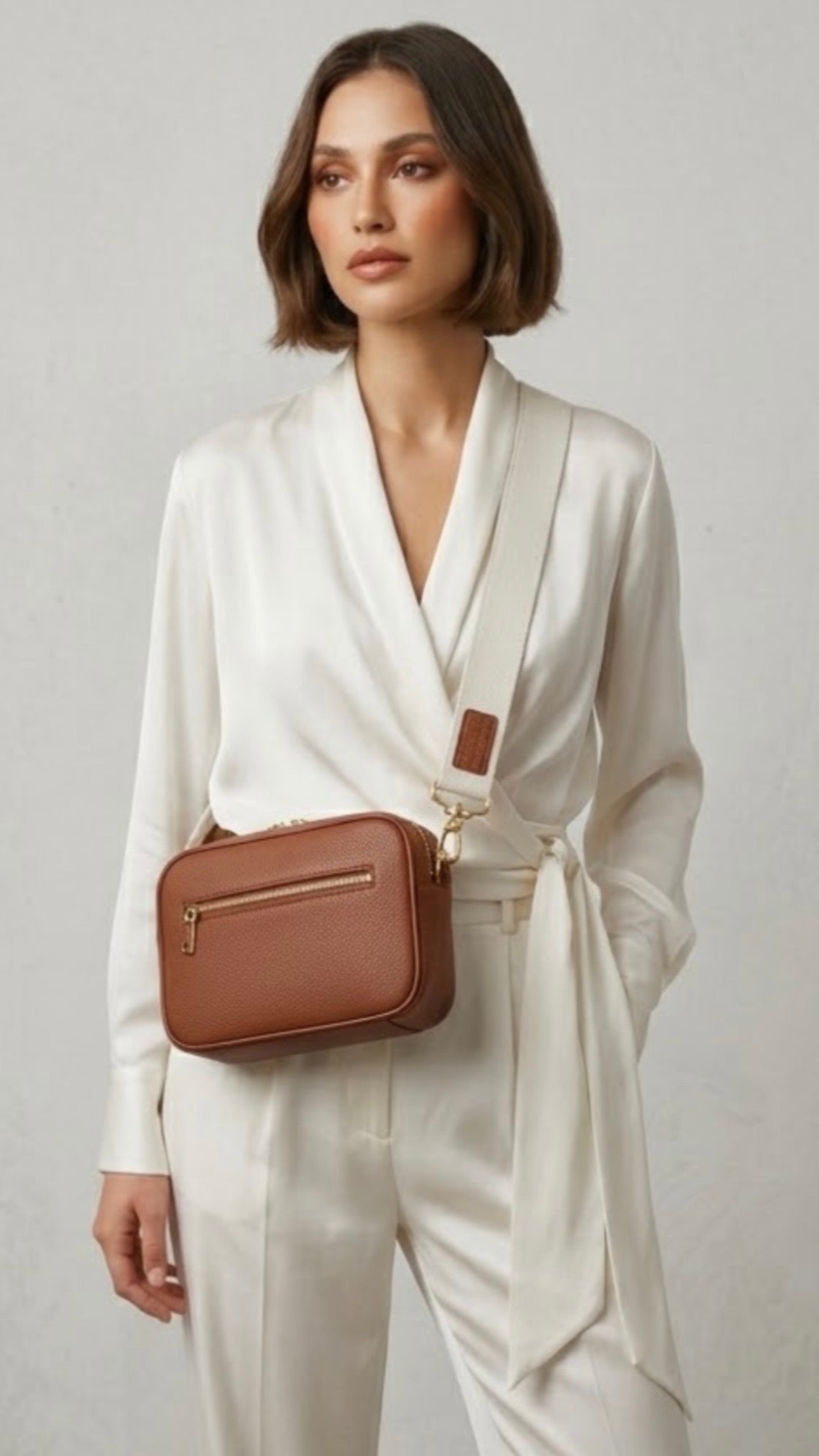 Classic elegance. TIKORI London classic pebbled leather crossbody bag in warm tan with adjustable strap and sleek design. Model in white silk cloth wears the cross body. Front profile of the bag shows in ivory background  Golden tone hardware and front profile of the bag with golden zipper detail. 