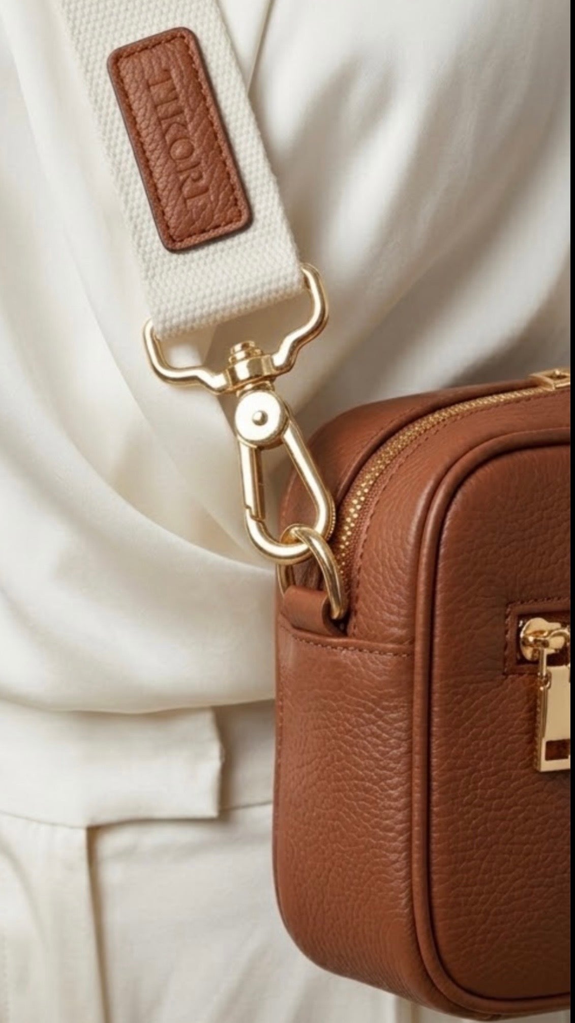 Classic elegance. TIKORI London classic pebbled leather crossbody bag in warm tan with adjustable strap and sleek design. Model in ivory cloths wears the crossbody. Close up details of the golden hardware and canvas strap with TIKORI logo showing in the picture. 