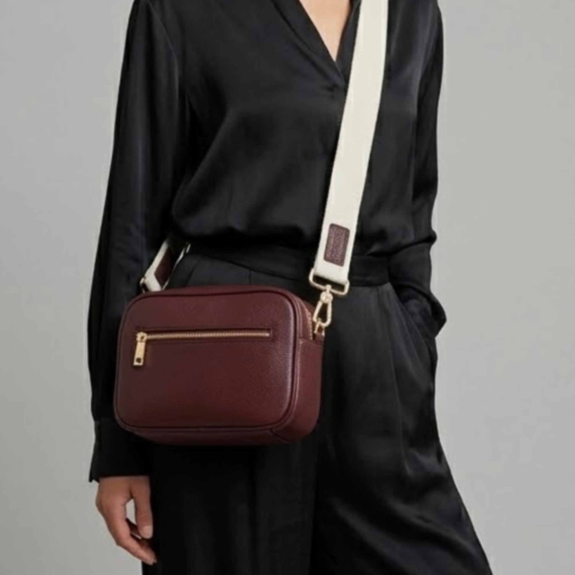Classic elegance with practicality and versatility in mind. TIKORI classic maroon by TIKORI london. Genuine Leather crossbody bag. Model in silk black cloths wears the crossbody . Close up detail of the bag of the bag showing with golden tone hardware. Middle zipper details and canvas strap with TIKORI logo shows in grey background. 