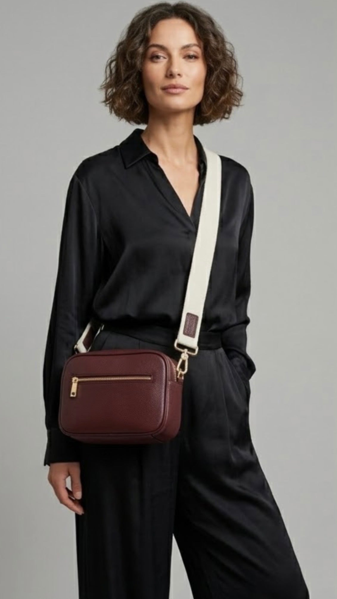 Classic elegance with practicality and versatility in mind. TIKORI classic maroon by TIKORI london. Genuine Leather crossbody bag. Model in black silk cloths wears the crossbody. Front profile of the bag showing with golden tone hardware. Front zipper details and front pocket shows in light grey background. 