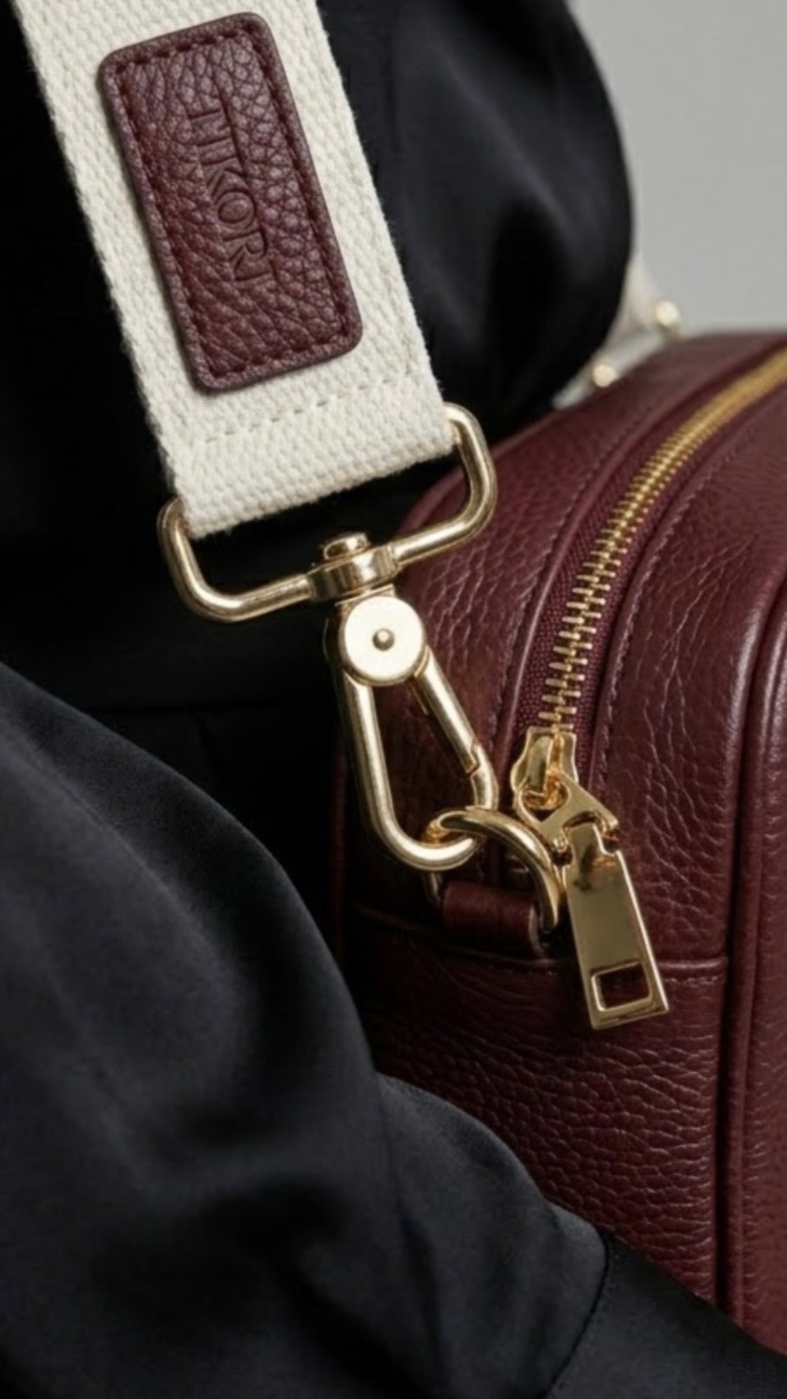 Classic elegance with practicality and versatility in mind. TIKORI classic maroon by TIKORI london. Genuine Leather crossbody bag. Close up detail of the bag of the bag showing with golden tone hardware. Middle zipper details and canvas strap with TIKORI logo shows in white background. 