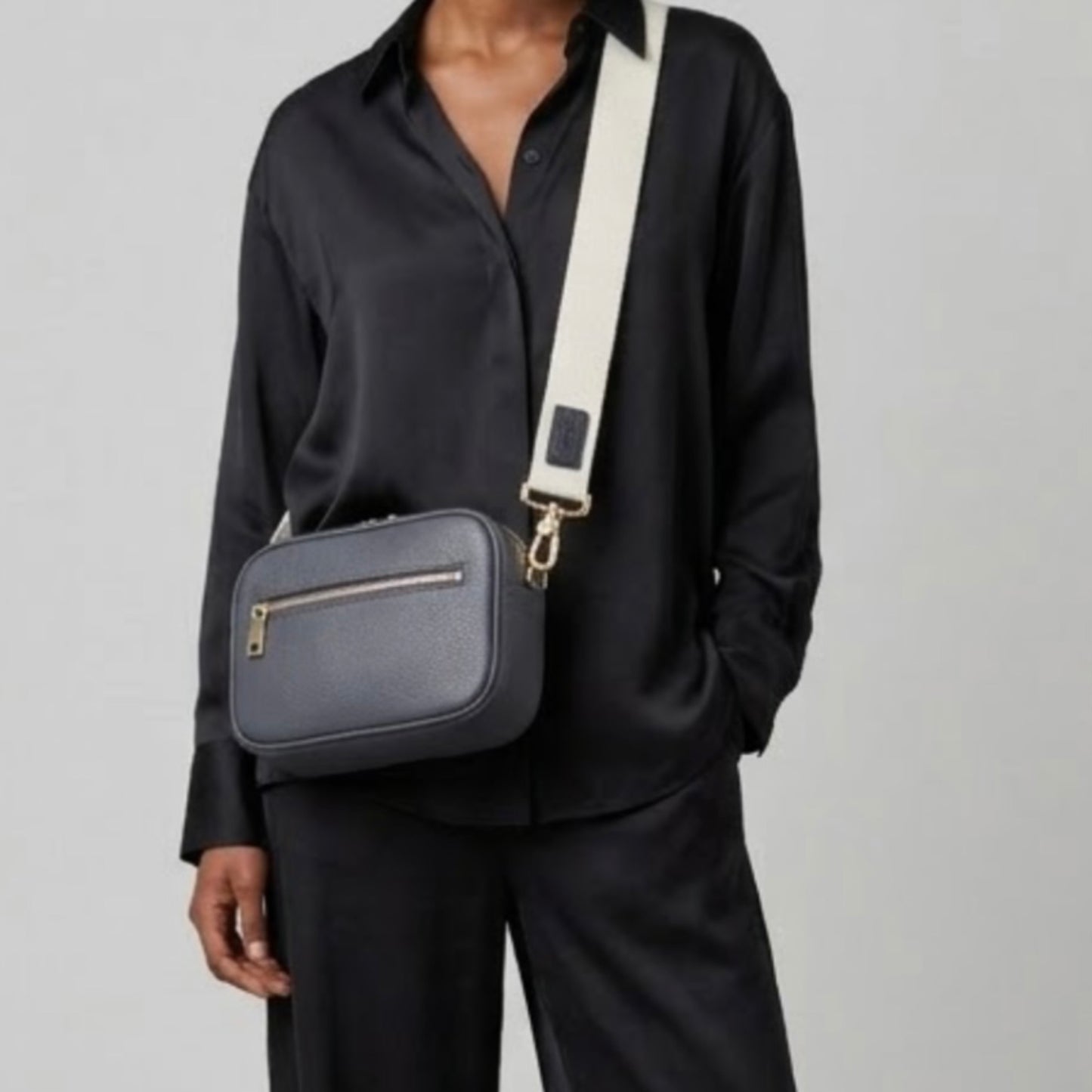 Classic elegance with practicality and versatility in mind. TIKORI classic grey purple by TIKORI London. Crossbody bag Crafted in soft pebbled premium genuine leather. Dark skinned beautiful Model body shows in the picture. She wears black silk cloths and wears the crossbody. Front profile of the bag showing golden hardware and front pocket with zipper details in white background  