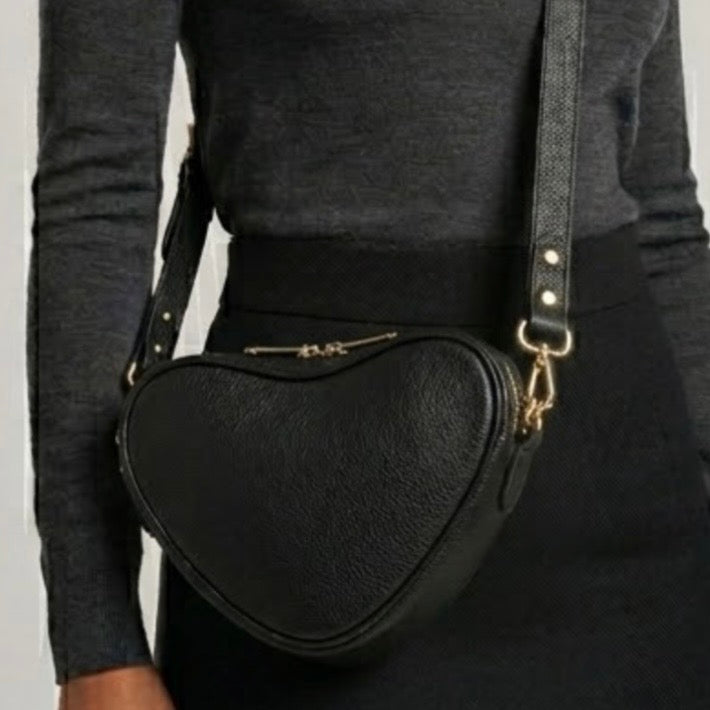 Selene heart crossbody bag by TIKORI London. Genuine premium leather with high quality craftsmanship and exquisite capacity design. Beautiful dark skinned body of a model in black tight shirt and dark grey top wears the crossbody. Long adjustable strap and golden tone hardware shows in soft grey natural colour background.