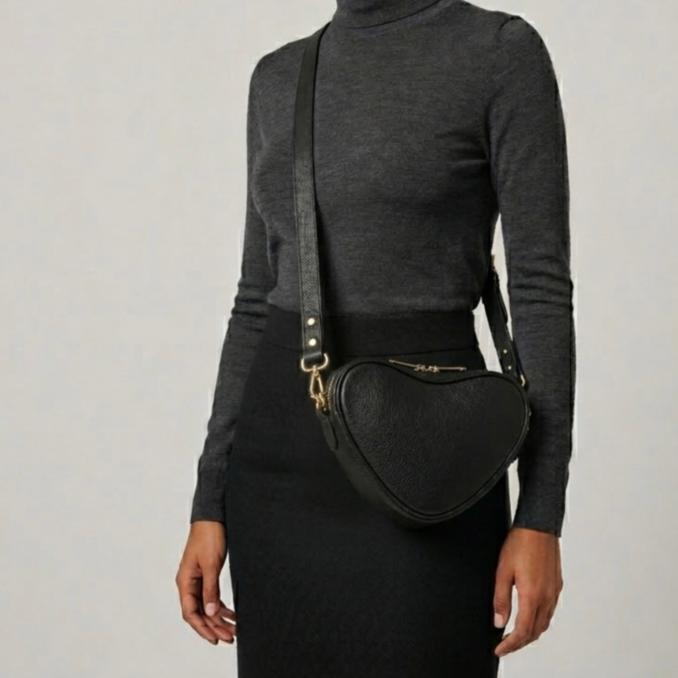 Selene heart crossbody bag by TIKORI London. Genuine premium leather with high quality craftsmanship and exquisite capacity design. Beautiful dark skinned body of a model in black tight shirt and dark grey top wears the crossbody facing right.  Long adjustable strap and golden tone hardware shows in soft grey natural colour background.