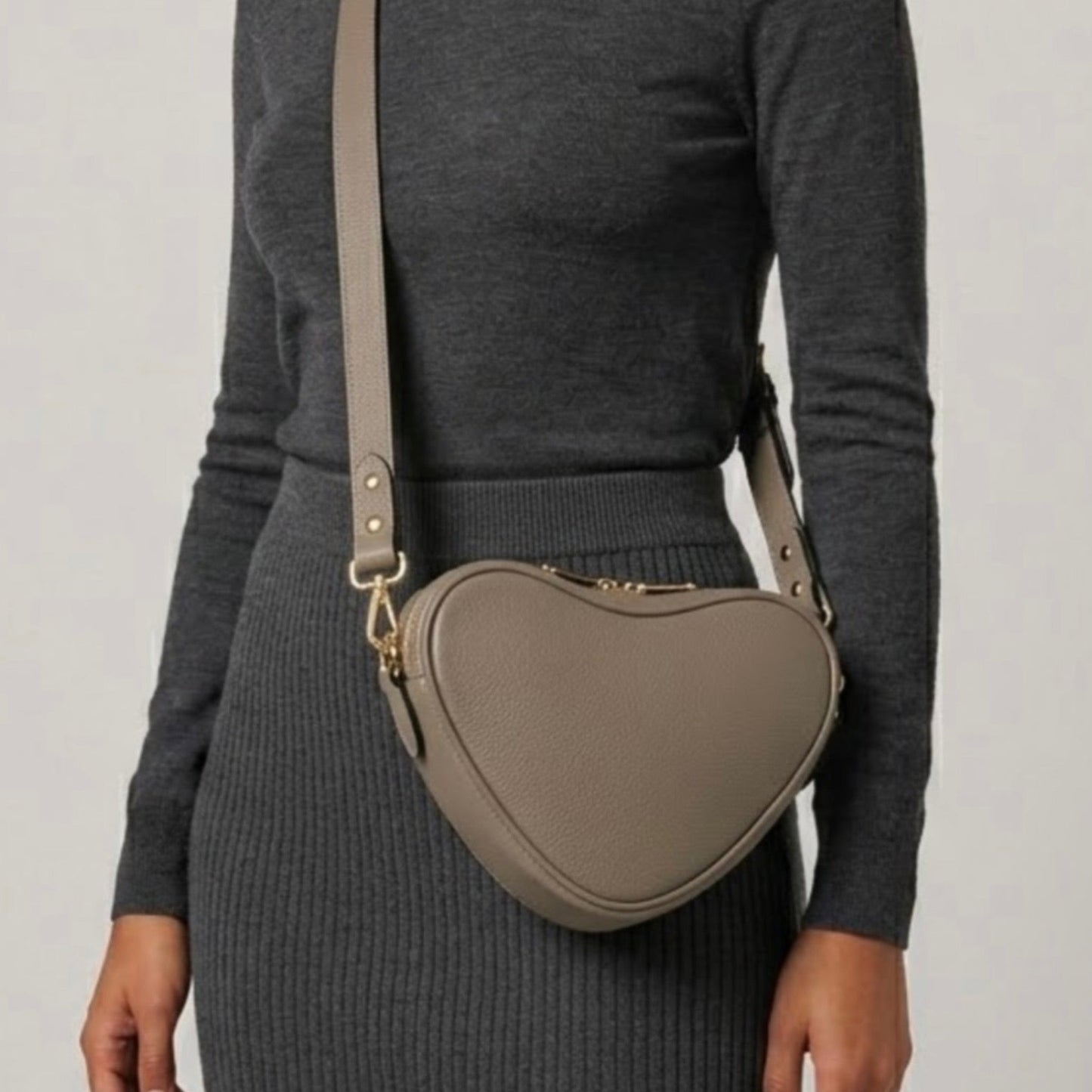 Beige heart-shaped leather crossbody bag by Tikori London, featuring a stylish and compact design. Body of a model wear the bag as a crossbody. She wears grey top and grey tight skirt . Bags long strap and shimmering golden hardware shows in a soft grey background.