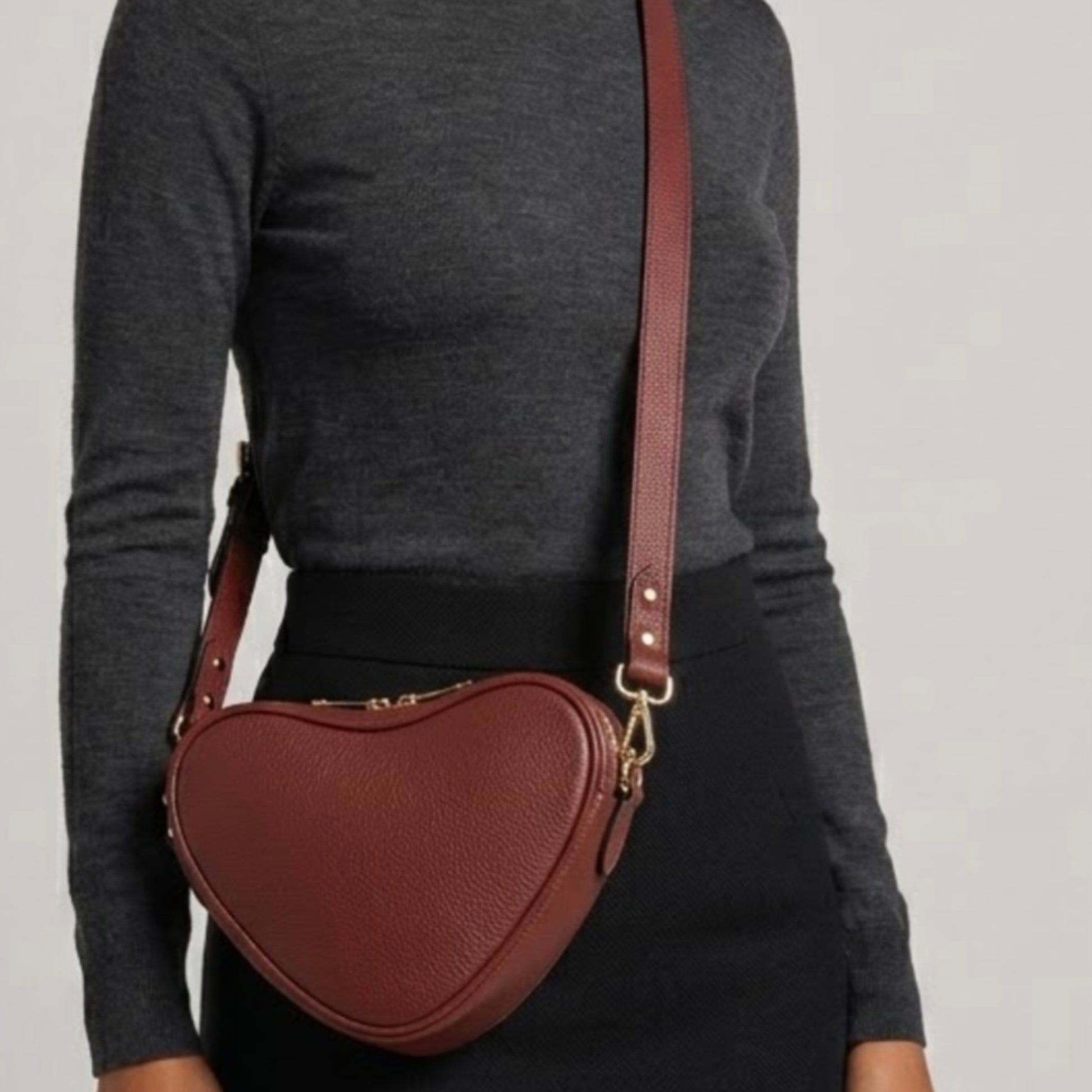 Red brown heart-shaped leather crossbody bag by Tikori London with adjustable strap and gold-tone hardware. Body of model wearing the bag as crossbody, showing in the picture. She wears dark grey top and black tight skirt in grey background. Bag and long strap prominently displayed.