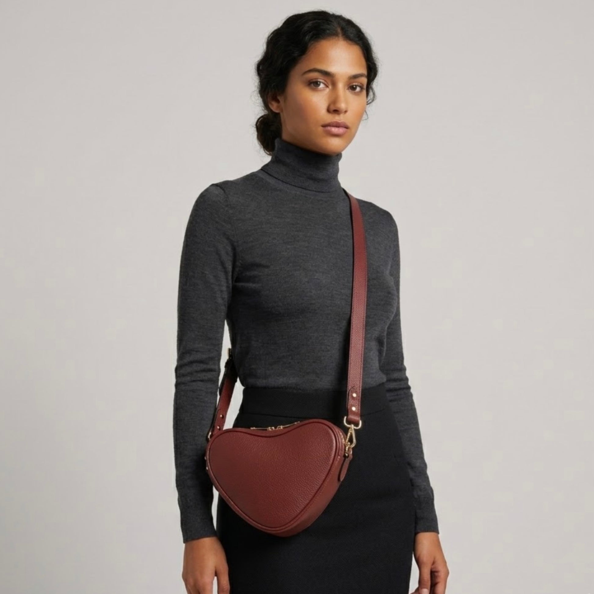 Red brown heart-shaped leather crossbody bag by Tikori London with adjustable strap and gold-tone hardware. Dark skinned beautiful model in black tight skirt and dark grey top wears the crossbody. Front profile of the bag showing in the picture with golden tone hardware and long strap highlighted in soft creamy background.