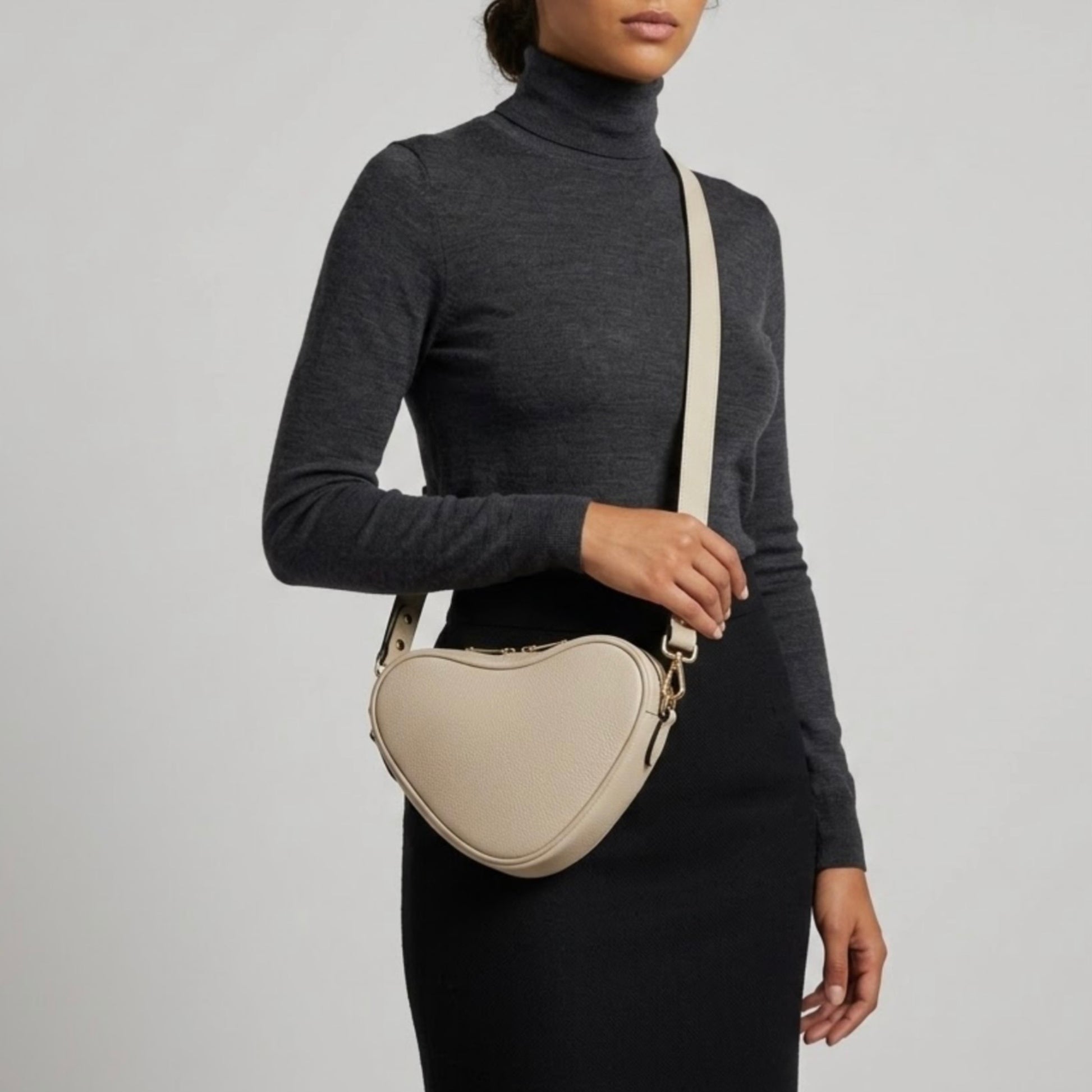 Selene heart-shaped leather crossbody bag Ivory by TIKORI  London with adjustable strap and gold-tone hardware. Dark skinned beautiful model in black tight skirt and dark grey top wears the crossbody. She is holding the strap from her left hand. Front profile of the bag showing in the picture with golden tone hardware and long strap highlighted in soft white  background.
