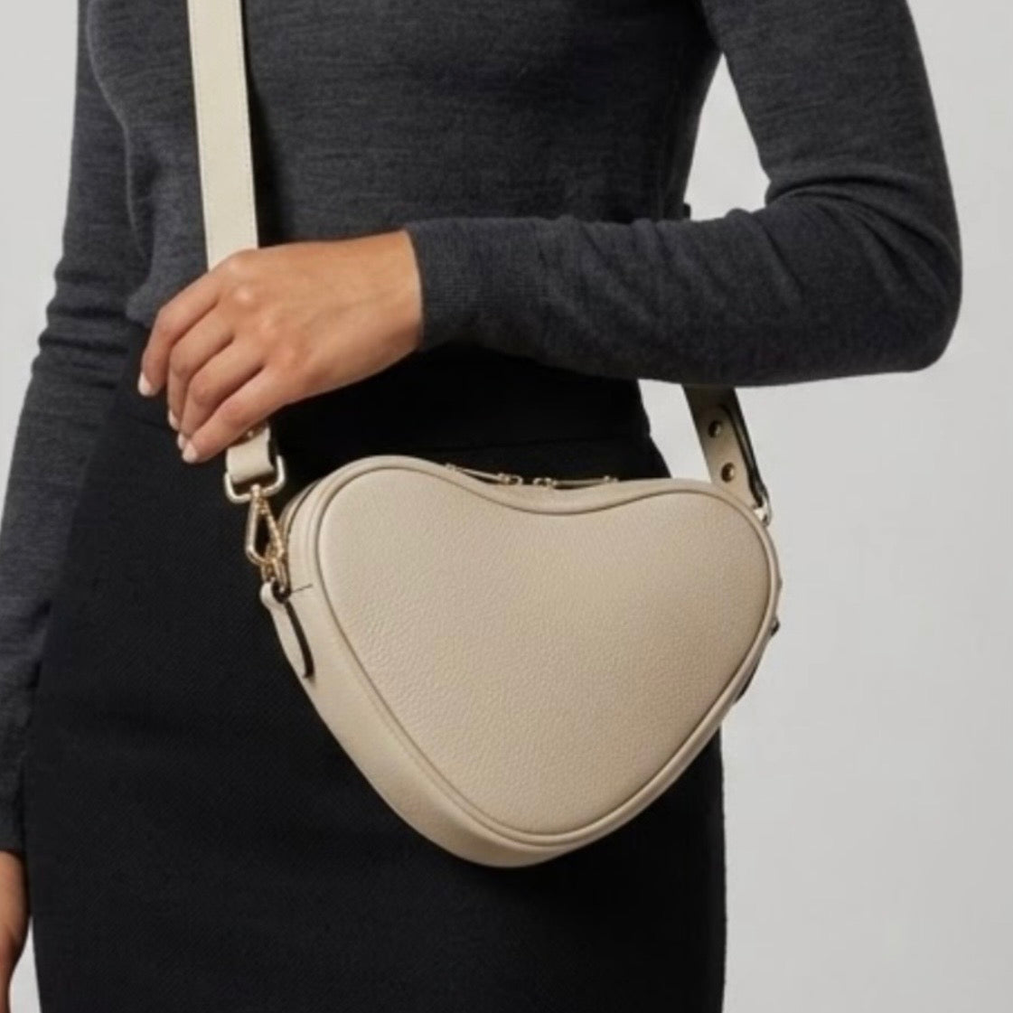 Selene heart-shaped leather crossbody bag Ivory by TIKORI  London with adjustable strap and gold-tone hardware. Dark skinned beautiful model in black tight skirt and dark grey top wears the crossbody. She is holding the strap from her left hand. Front profile of the bag showing in the picture with golden tone hardware and long strap highlighted in soft white  background.