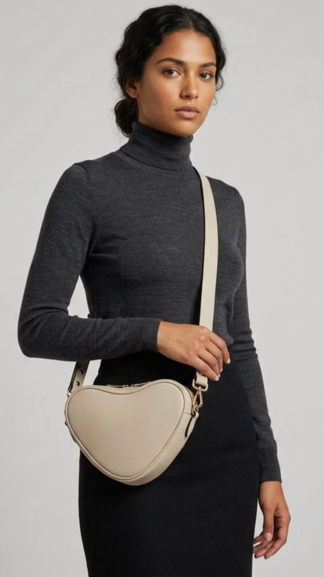 Selene heart-shaped leather crossbody bag Ivory by TIKORI  London with adjustable strap and gold-tone hardware. Full picture of a Dark skinned beautiful model in black tight skirt and dark grey top wears the crossbody. She is holding the strap from her left hand. Front profile of the bag showing in the picture with golden tone hardware and long strap highlighted in soft white  background.