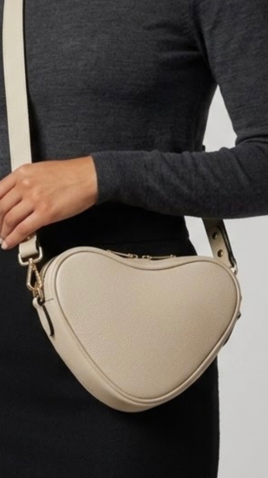 Selene heart-shaped leather crossbody bag Ivory by TIKORI  London with adjustable strap and gold-tone hardware. Dark skinned beautiful model in black tight skirt and dark grey top wears the crossbody. She is holding the strap from her left hand. Close up detail of the bag showing in the picture with golden tone hardware and long strap highlighted in soft white  background.