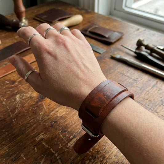 Dark brown leather layered bracelet by TIKORI London with smooth leather layered look, stylish accessory for men and women. Male model hand displays wearing the dark brown bracelet. Table with leather items and tools also shows in the picture. Leather workshop environment.