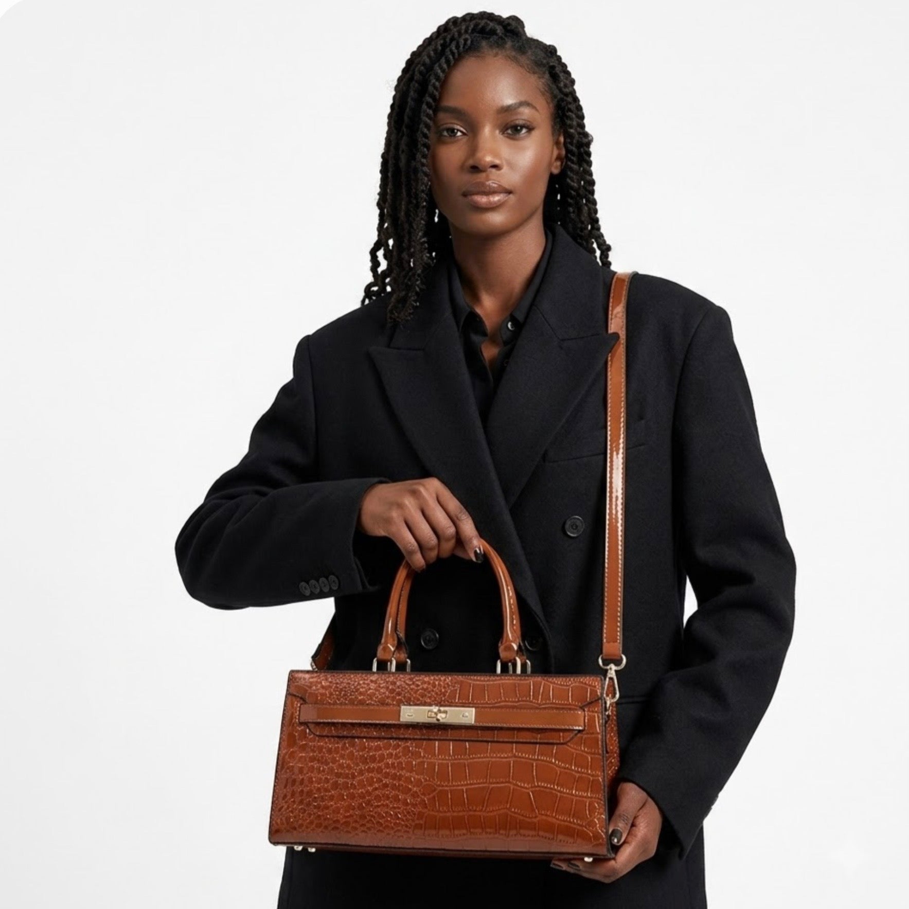 Gilded Croc Baguette Shoulder Bag cognac by TIKORI LONDON. Designed with a structured architectural frame and a glossy crocodile-embossed finish, it brings a "quiet luxury" aesthetic to your wardrobe at a fraction of the cost. Beautiful dark skinned model in black oversized coat wearing the bag as cross body. Front profile showing with golden front lock in a fully white background.