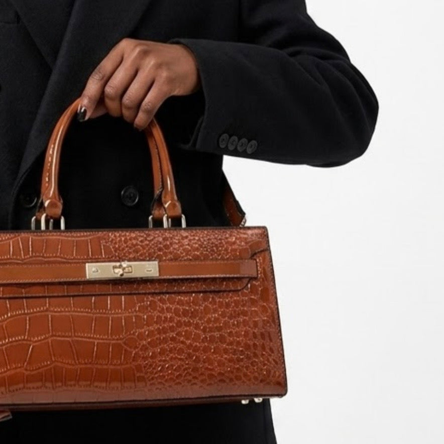 Gilded Croc Baguette Shoulder Bag cognac by TIKORI LONDON. Designed with a structured architectural frame and a glossy crocodile-embossed finish, it brings a "quiet luxury" aesthetic to your wardrobe at a fraction of the cost. Beautiful dark skinned model in black oversized coat holding the bag from top handle. Middle part of the model body can be seen in fully white background.