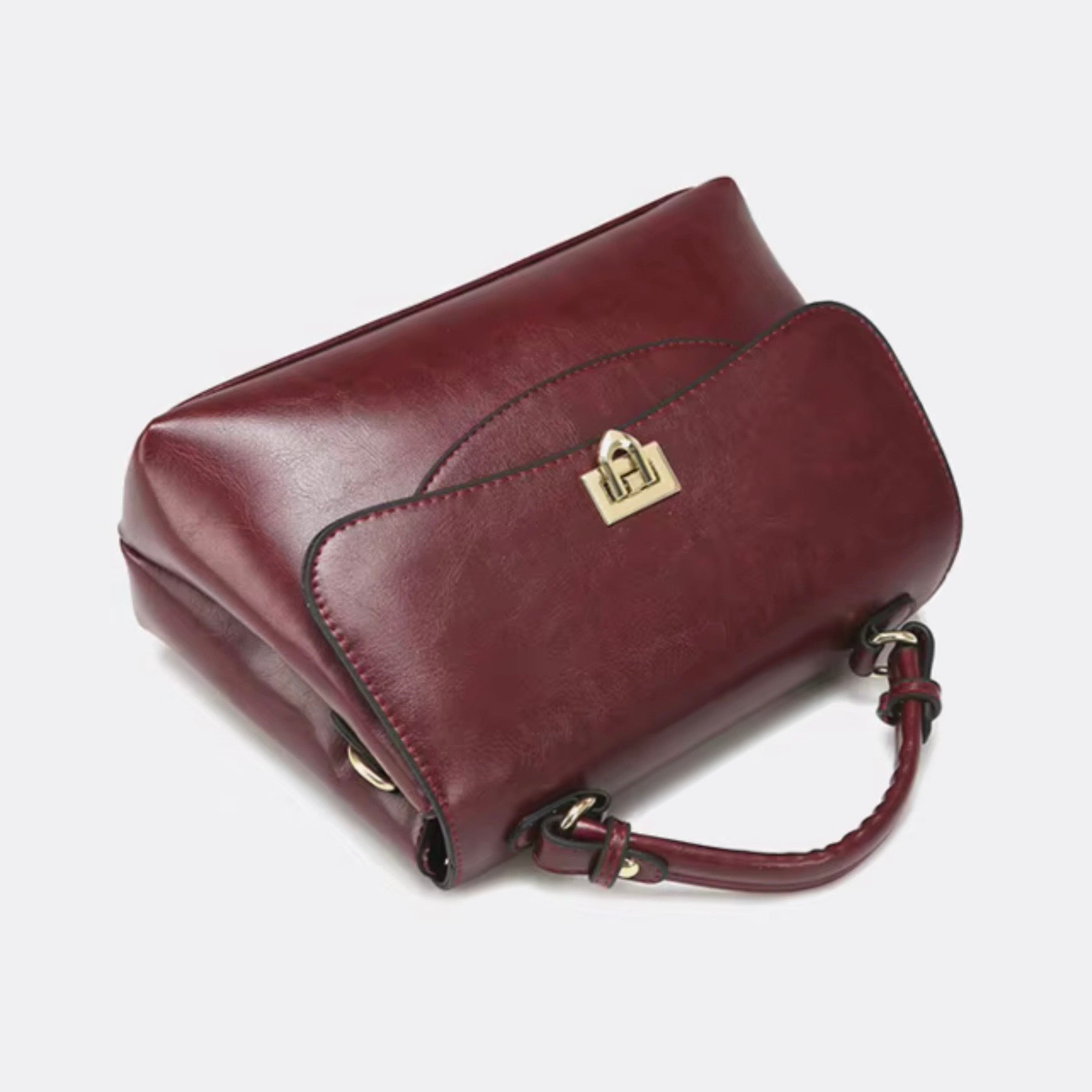 Heritage flap bag maroon by TIKORI London. Defined by a unique braided leather handle that adds a touch of luxury. Features a beautiful, sweeping front flap secured by a polished, shield shaped gold clasp. Bag laid on the floor showing the front profile. Golden lock in the middle and curved top handle beautifully display in a fully white background.