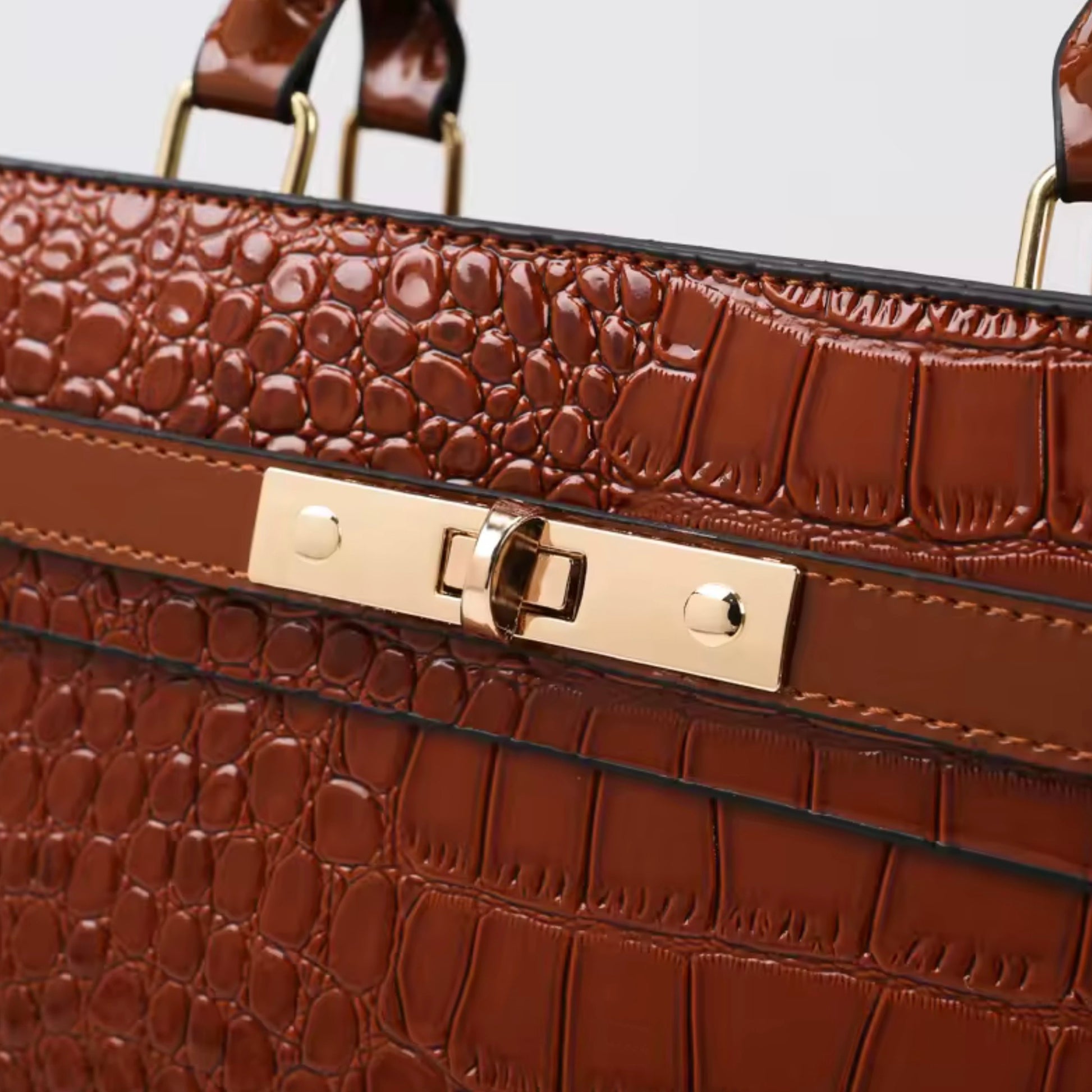 Gilded Croc Baguette Shoulder Bag cognac by TIKORI LONDON. Designed with a structured architectural frame and a glossy crocodile-embossed finish, it brings a "quiet luxury" aesthetic to your wardrobe at a fraction of the cost. Close up detail of the bags front golden hardware lock showing in a fully white background.