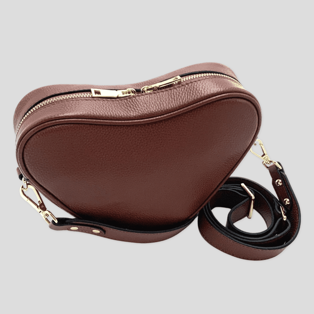 Red brown leather heart-shaped crossbody bag by Tikori London, featuring a compact design with a strap and stylish finish. Front profile of the bag. Showing thick long strap with golden hardware and zipper detail shows in soft light grey background.