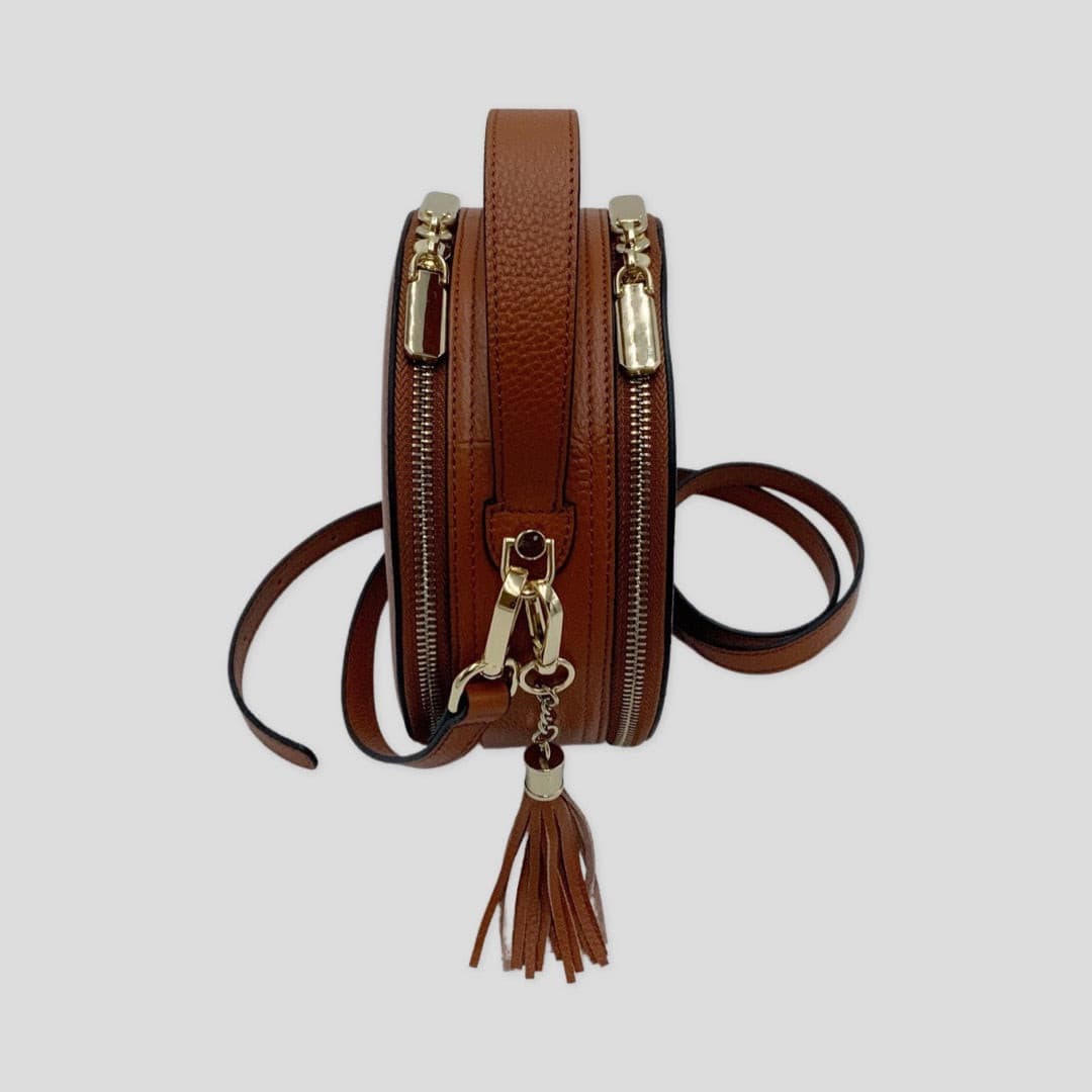 Cleopatra by TIKORI London. warm tan leather circle crossbody bag with dual zip fastening and finished leather design. Side profile of the bag showing in the picture.