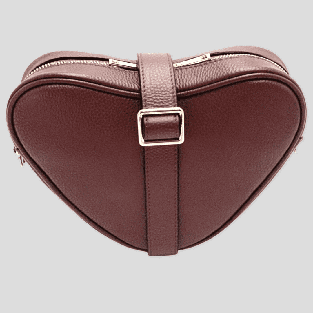 Red brown heart-shaped leather crossbody bag by Tikori London with adjustable strap and gold-tone hardware. Front profile of the bag showing in the picture. Strap is wrapped around the bag showing the golden tone buckle in soft grey background.