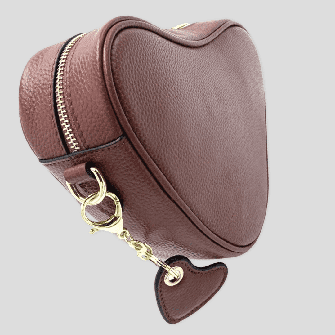 Red brown leather heart-shaped crossbody bag by Tikori London, featuring a compact design and adjustable strap. Side profile of the bag showing golden hardware and heart shaped tussle hanging from the side of the bag. Displayed in soft grey background 